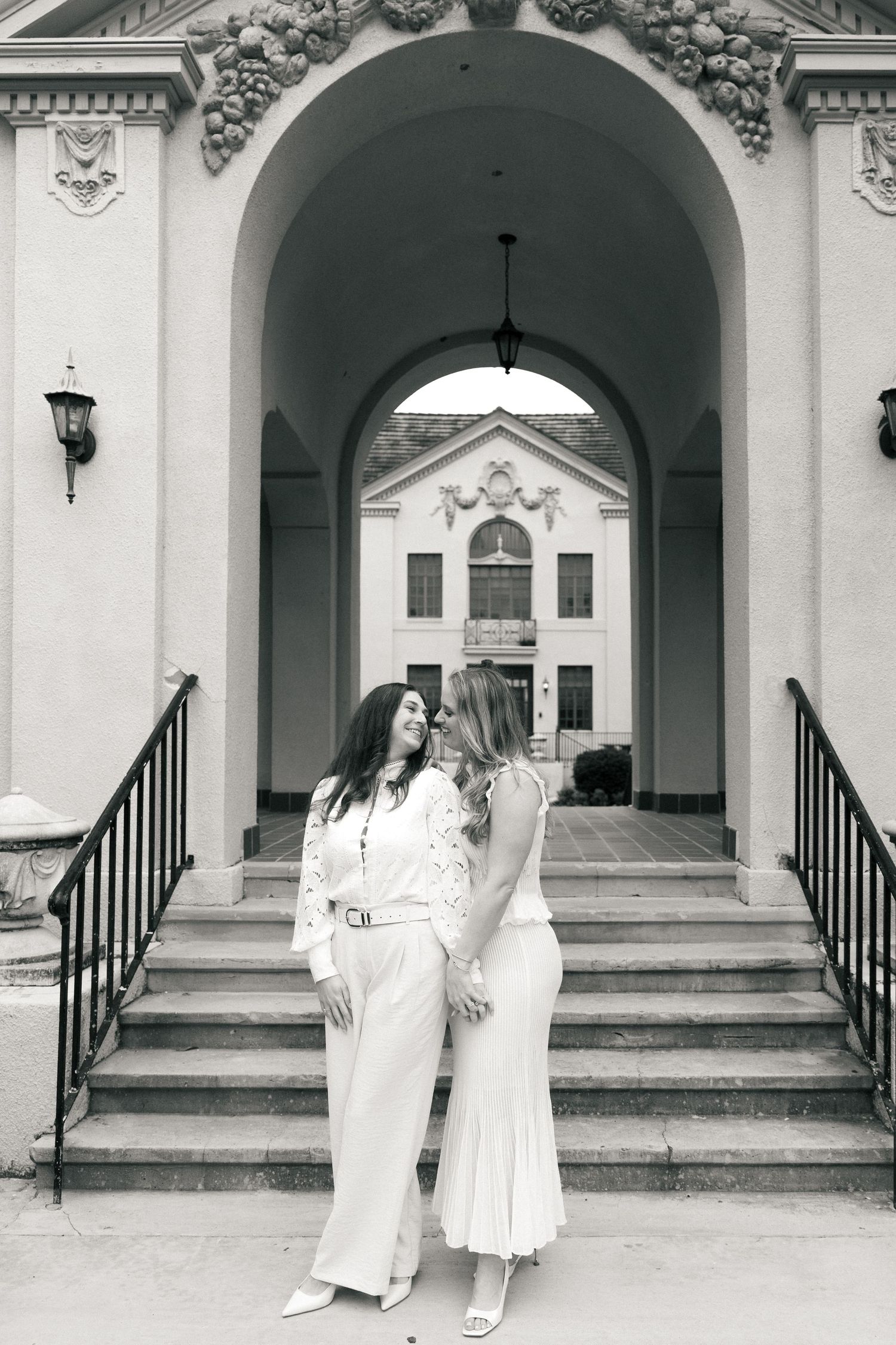 Two women holding hands in white outfits before an elegant building with arched windows and ornate details.