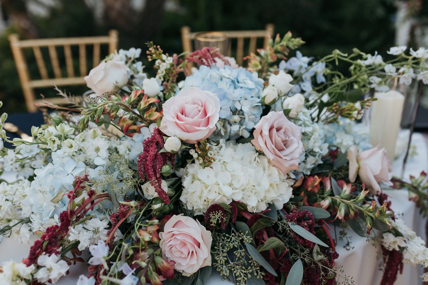 Elegant wedding centerpiece with blush pink roses, white hydrangeas, dusty blue blooms, and burgundy accents on a reception table.