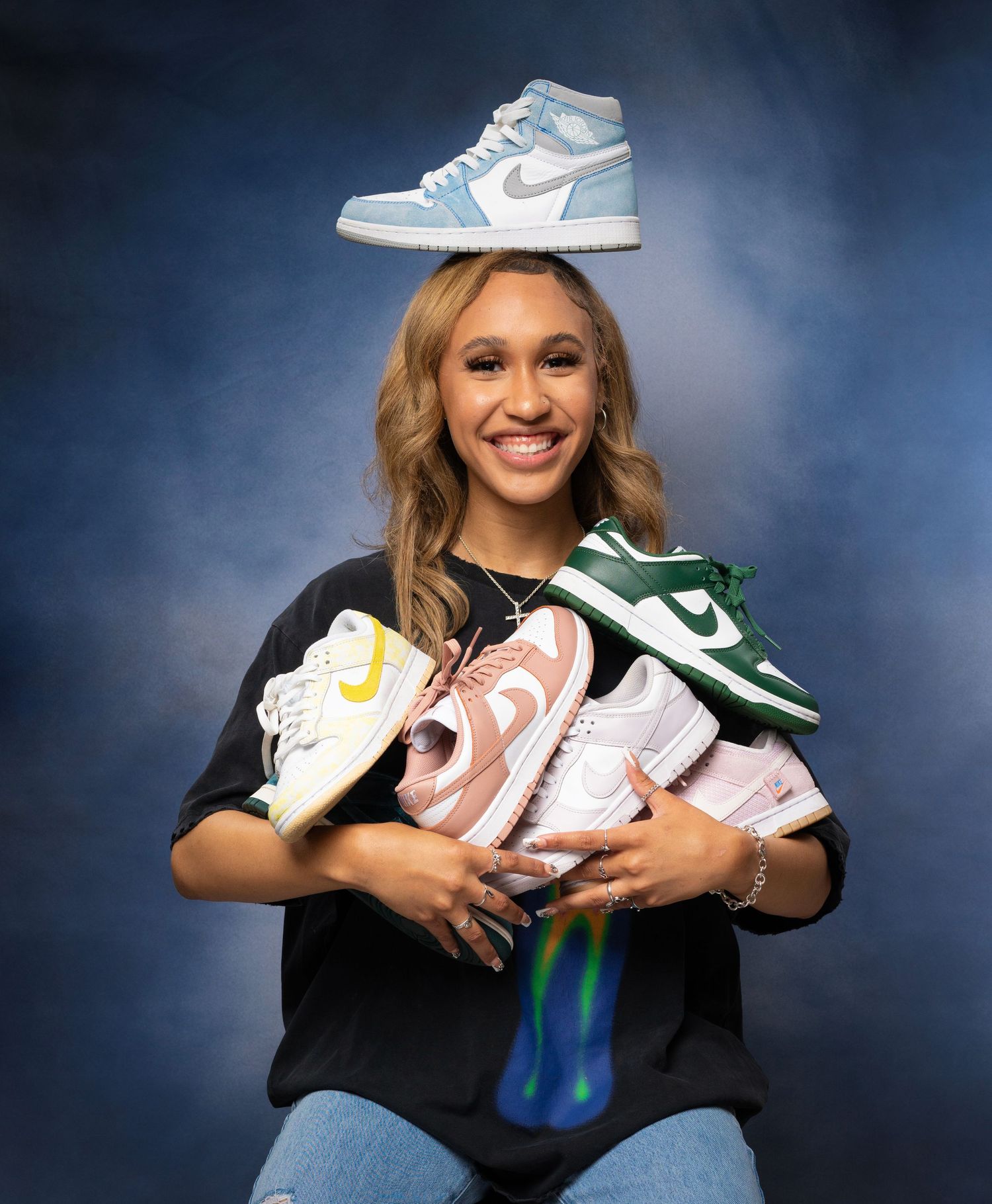 Smiling young woman holds collection of Nike sneakers including Jordan 1s and Dunks in studio.