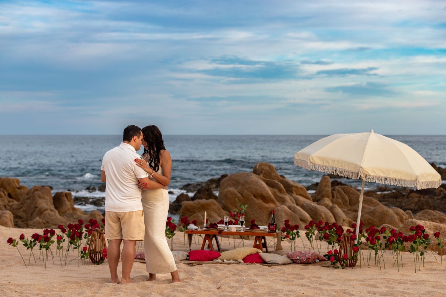 Romantic couple embracing on a sandy beach surrounded by red roses and a white umbrella at sunset.