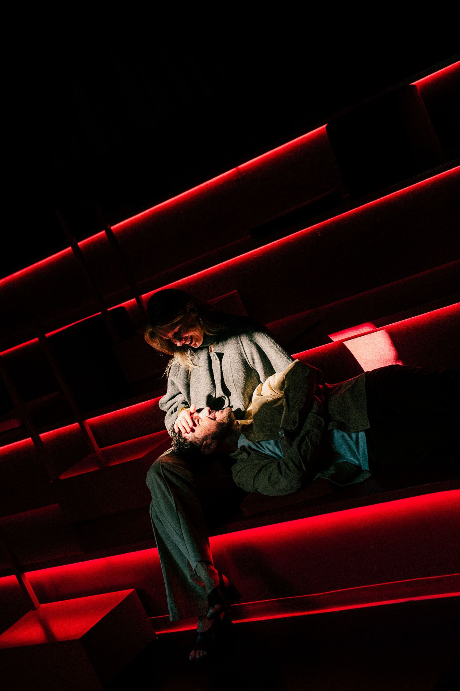Two performers in dramatic red-lit theater scene, one cradling the other on stepped stage risers.