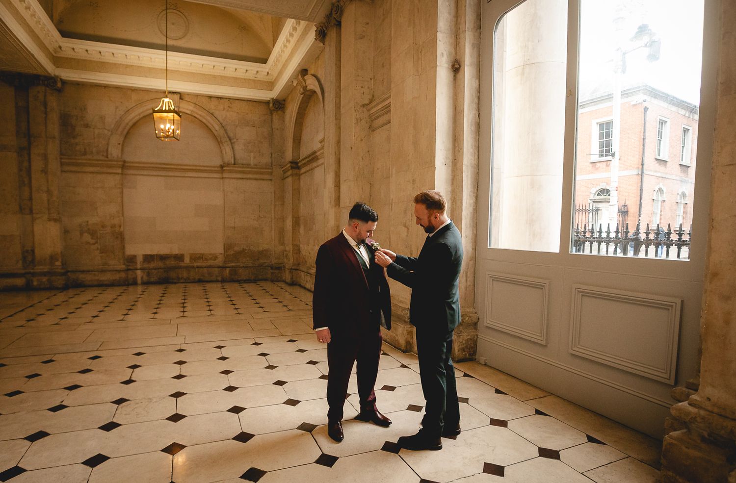 Two grooms in suits share a tender moment in an elegant marble-floored hall during their wedding day.