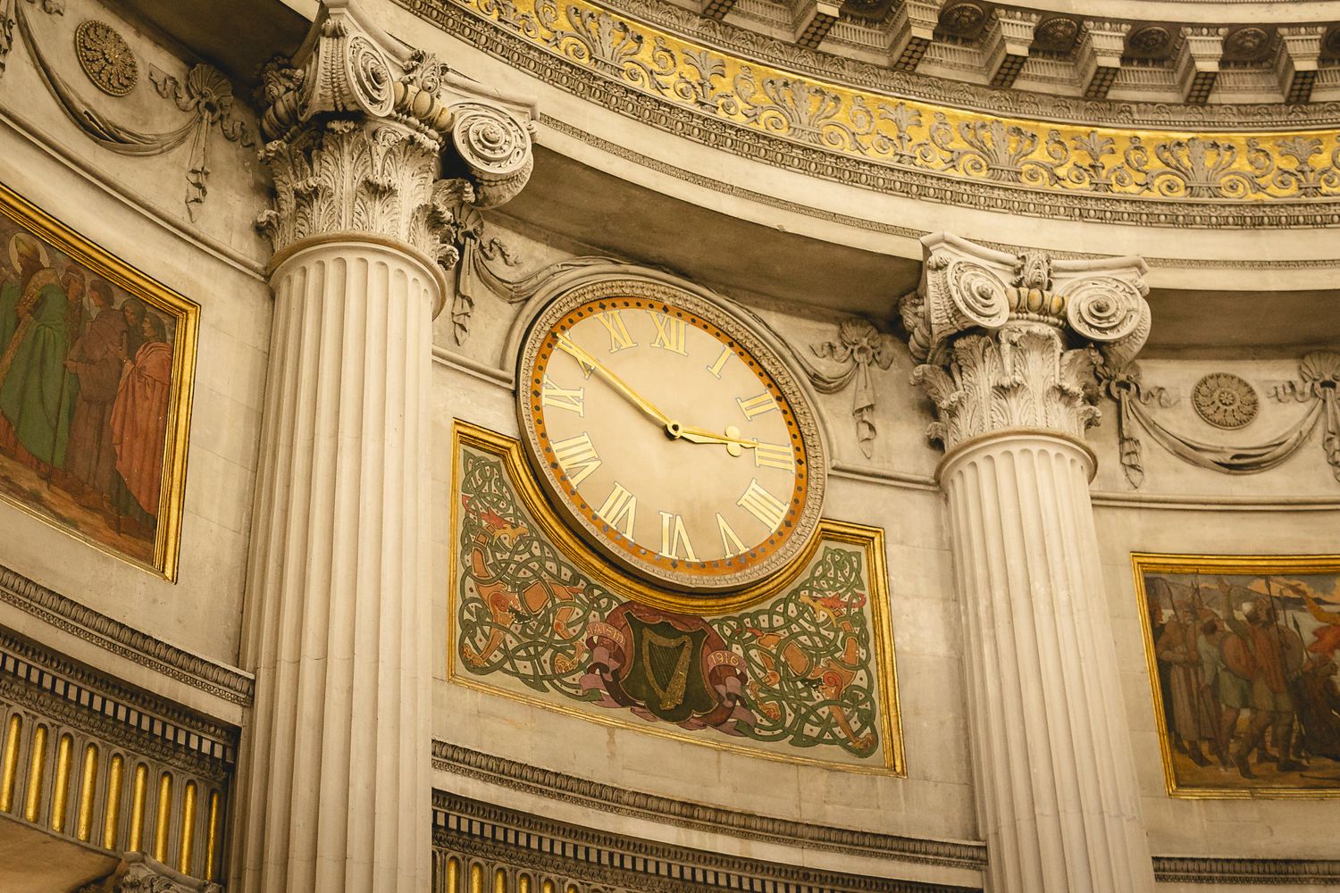 Ornate clock with Roman numerals on a neoclassical building interior with gilded details and Corinthian columns.