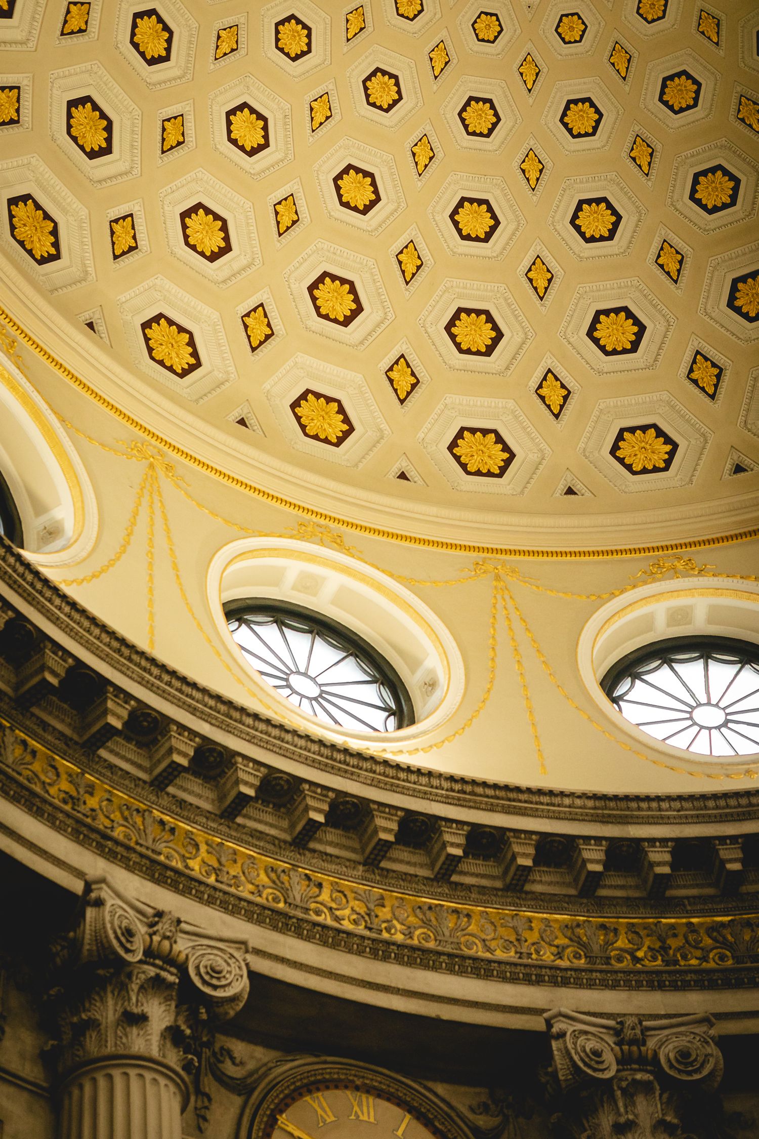 Interior view of a neoclassical dome ceiling with gold hexagonal coffers and circular stained glass windows.