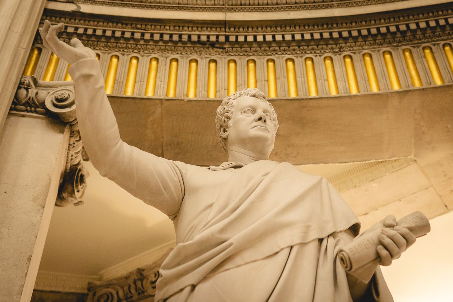 Classical marble statue of a robed figure with raised arm inside a neoclassical building with ornate columns.