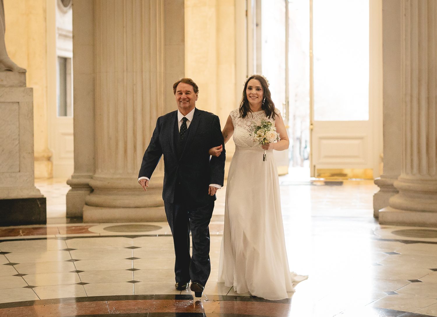 Bride in white gown walks with escort through grand columned hall on wedding day.
