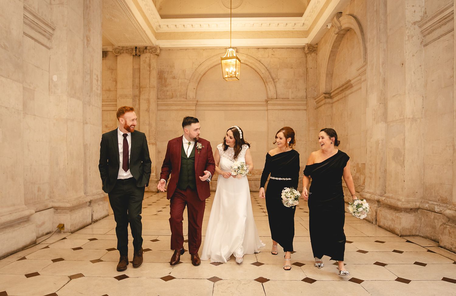 Wedding party walking together in elegant marble hallway, bride in white gown with groom in burgundy suit and guests in formal attire.