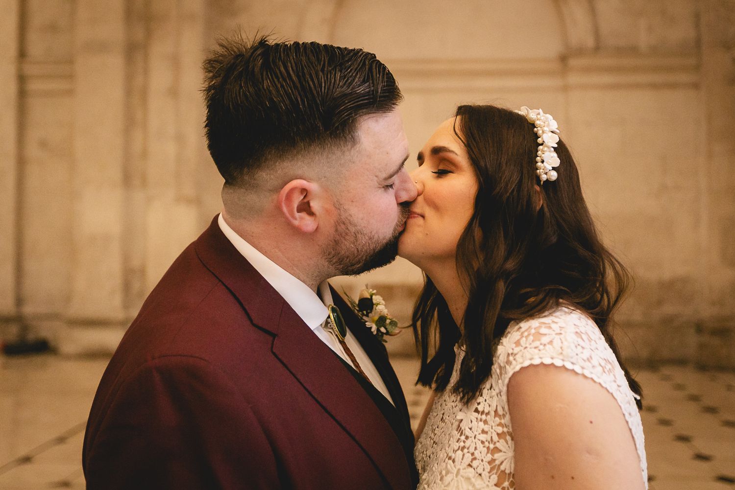 Bride and groom sharing a romantic kiss at their wedding, groom in burgundy suit, bride in lace dress with floral hair piece.