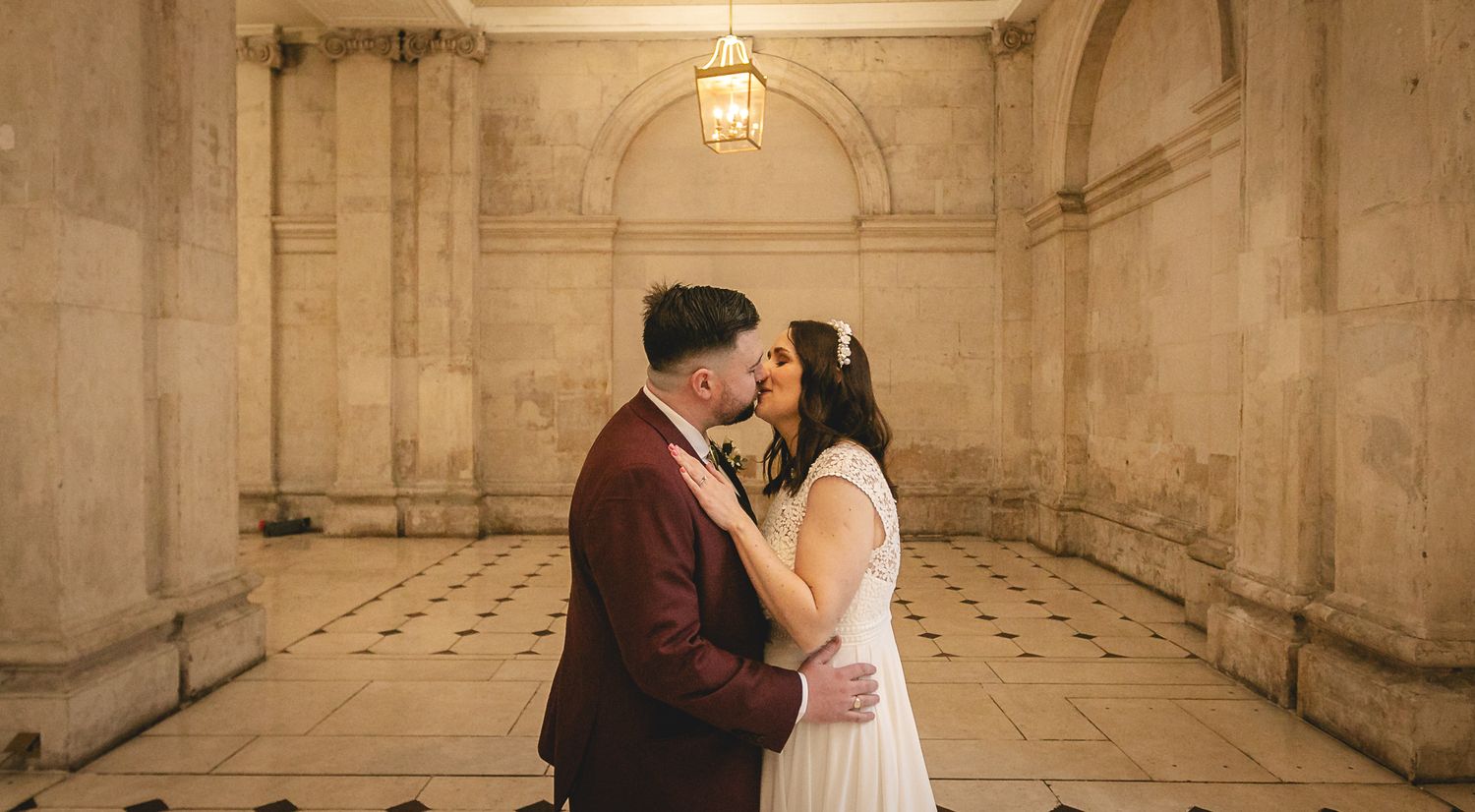 Bride and groom sharing a romantic kiss in a warmly lit stone archway alcove during their wedding day.
