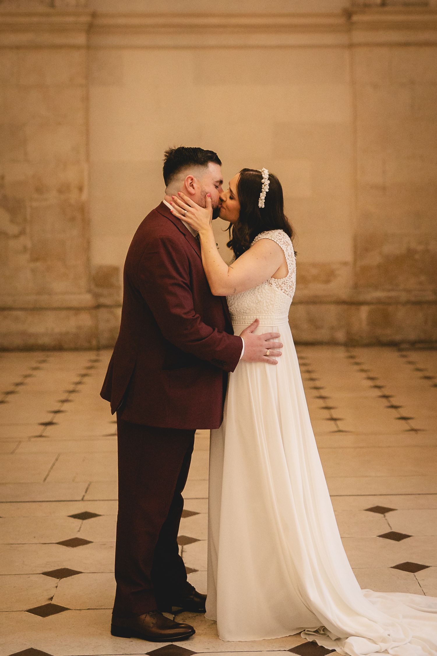 Newlyweds share first kiss in elegant marble-floored venue with groom in burgundy suit.