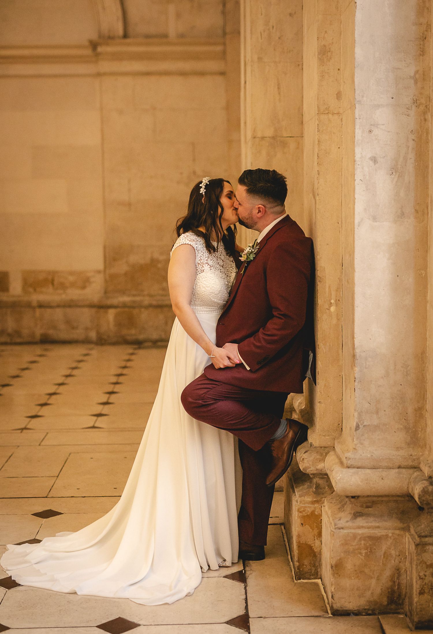 Bride and groom sharing a romantic kiss against a stone column in an elegant historic venue.