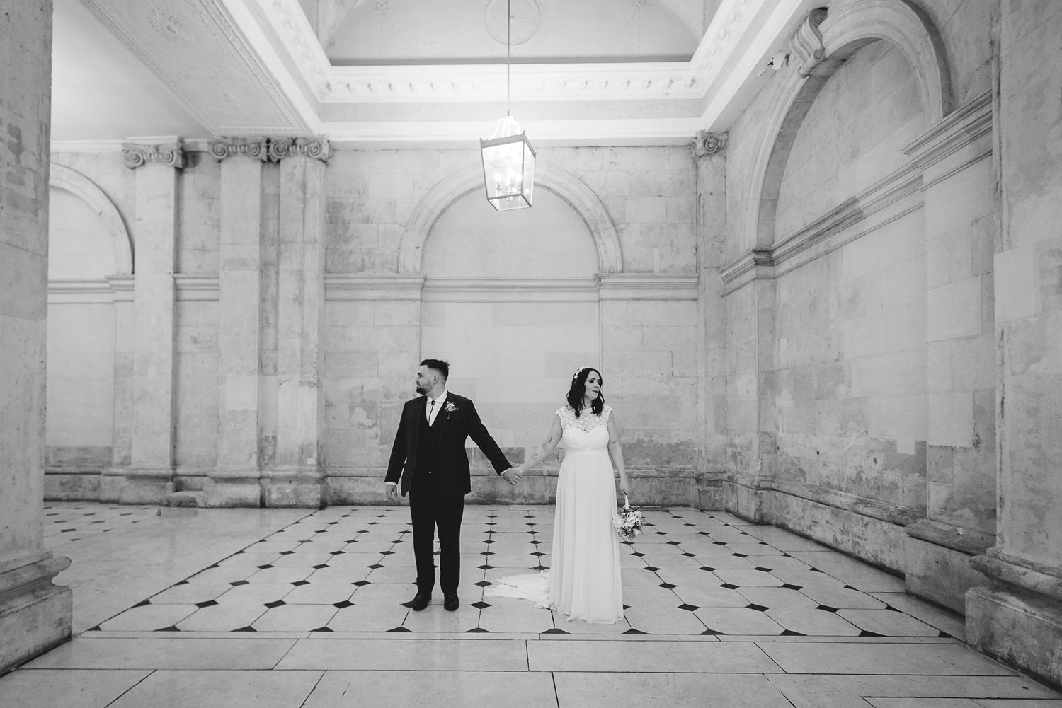 Bride and groom hold hands in a grand marble hall with checkered floors in black and white photo.