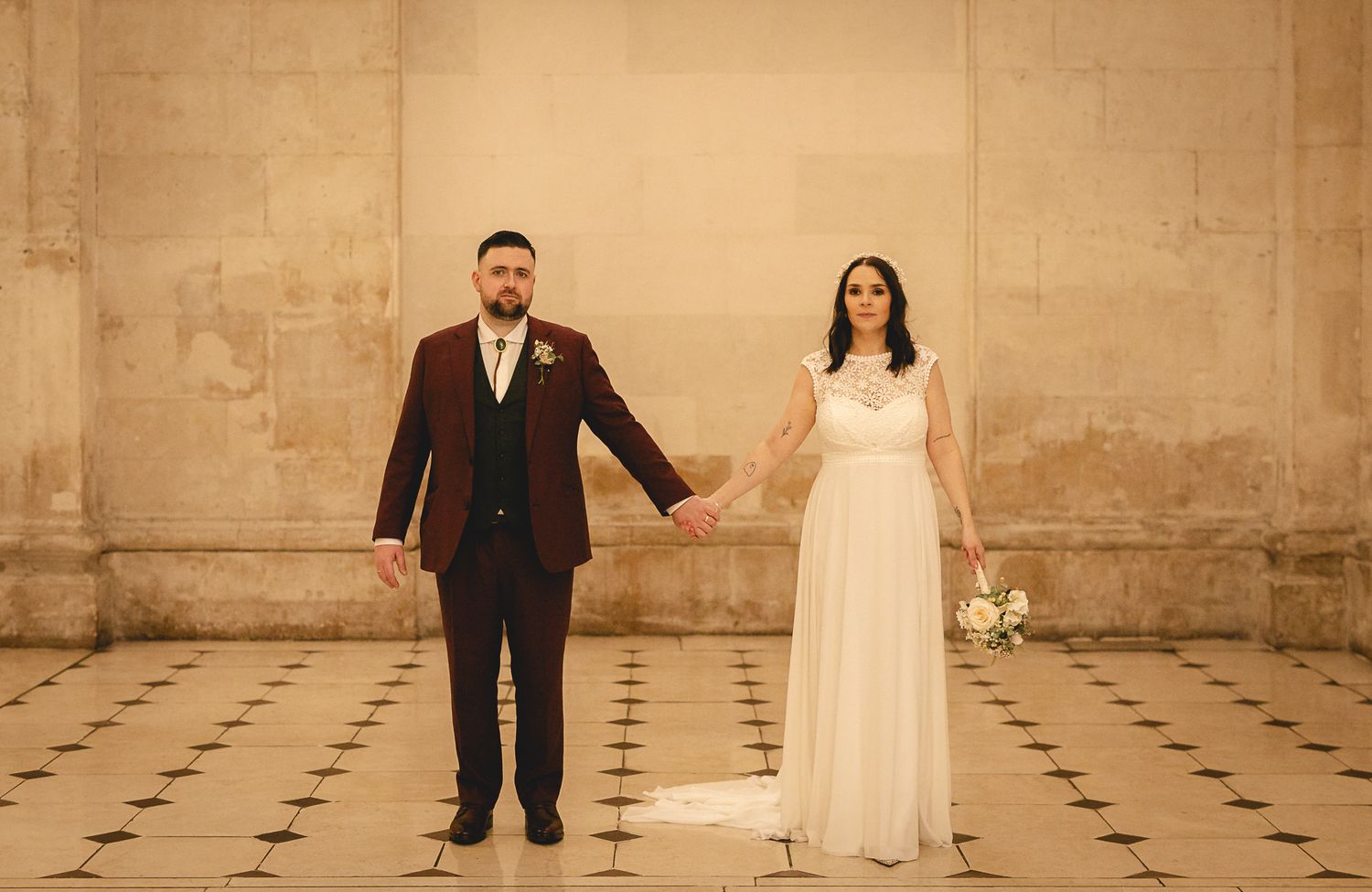 Bride and groom holding hands in elegant venue, groom in burgundy suit, bride in white flowing gown holding bouquet.