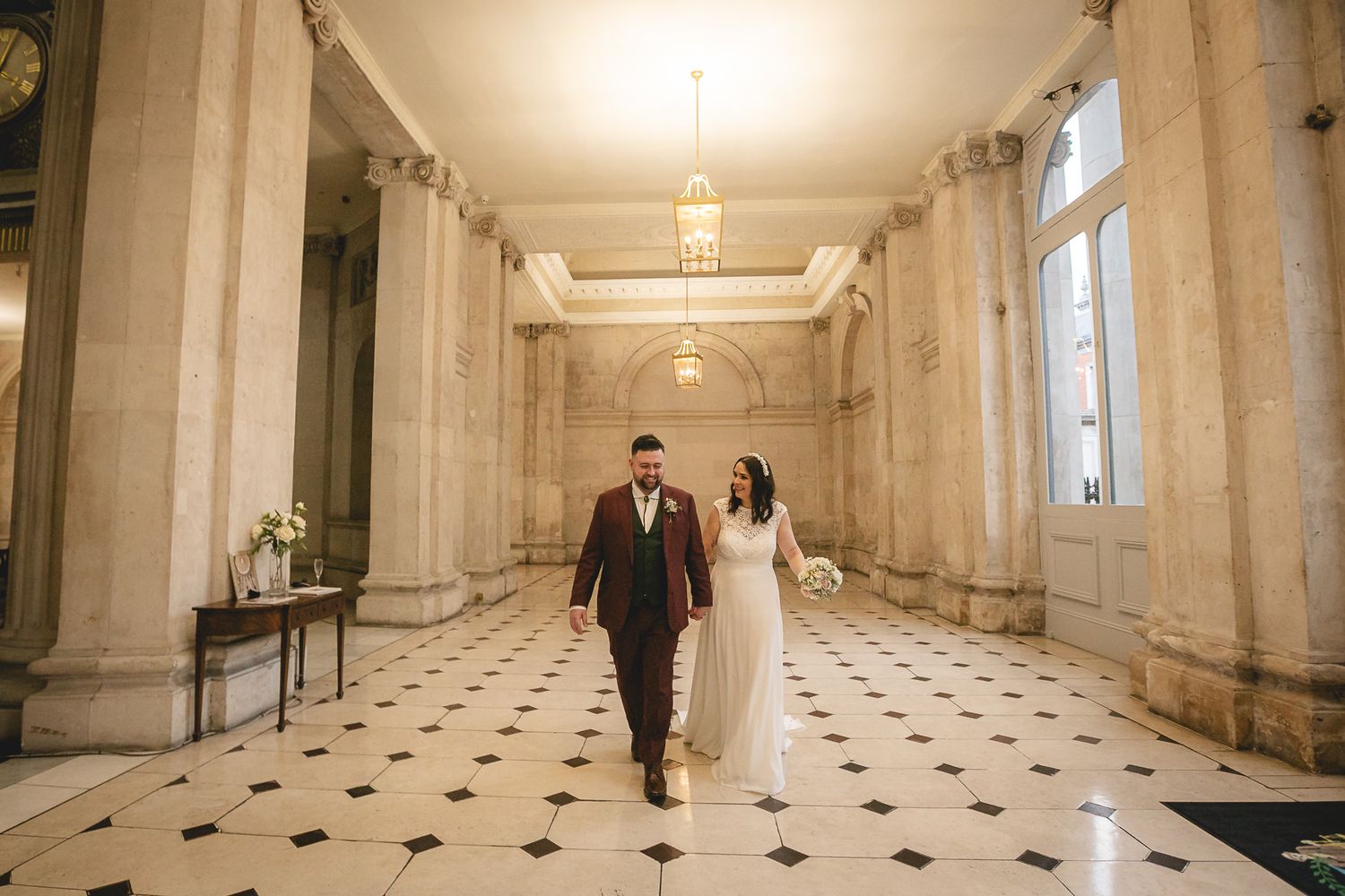 Bride and groom walking hand in hand through an elegant marble-columned hallway with black and white checkered floors.