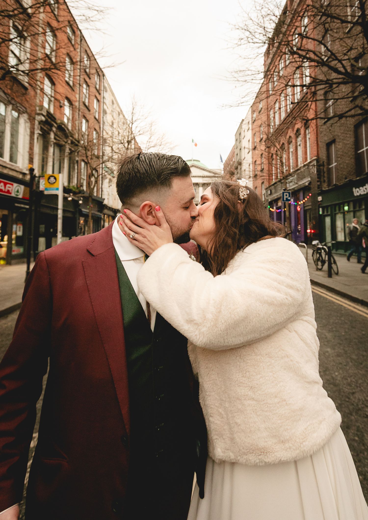 Newlyweds kiss on bustling city street, bride in white fur coat and groom in burgundy suit.