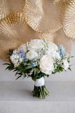A beautiful bridal bouquet featuring white roses and light blue hydrangeas tied with a white ribbon against a textured backdrop.