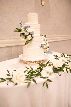 Elegant white tiered wedding cake decorated with gentle blue and white flowers and greenery on a gold cake stand.