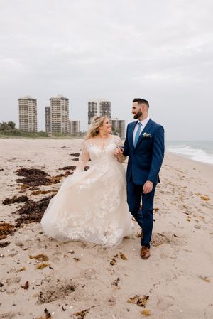 Newlyweds share a romantic moment walking along a beach with high-rise buildings in the background on an overcast day.