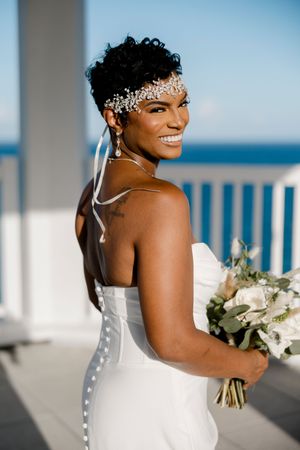 Beautiful bride in a white strapless wedding dress holds a white bouquet on a beachfront balcony.