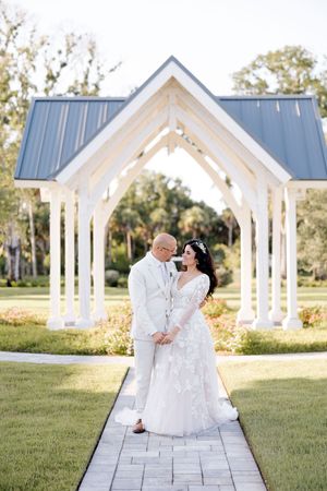 A romantic wedding couple sits and walks along a white pavilion pathway in an outdoor garden setting.