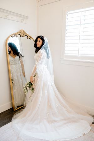 A beautiful white wedding dress with a long train is displayed beside an ornate gold mirror in a bright room.
