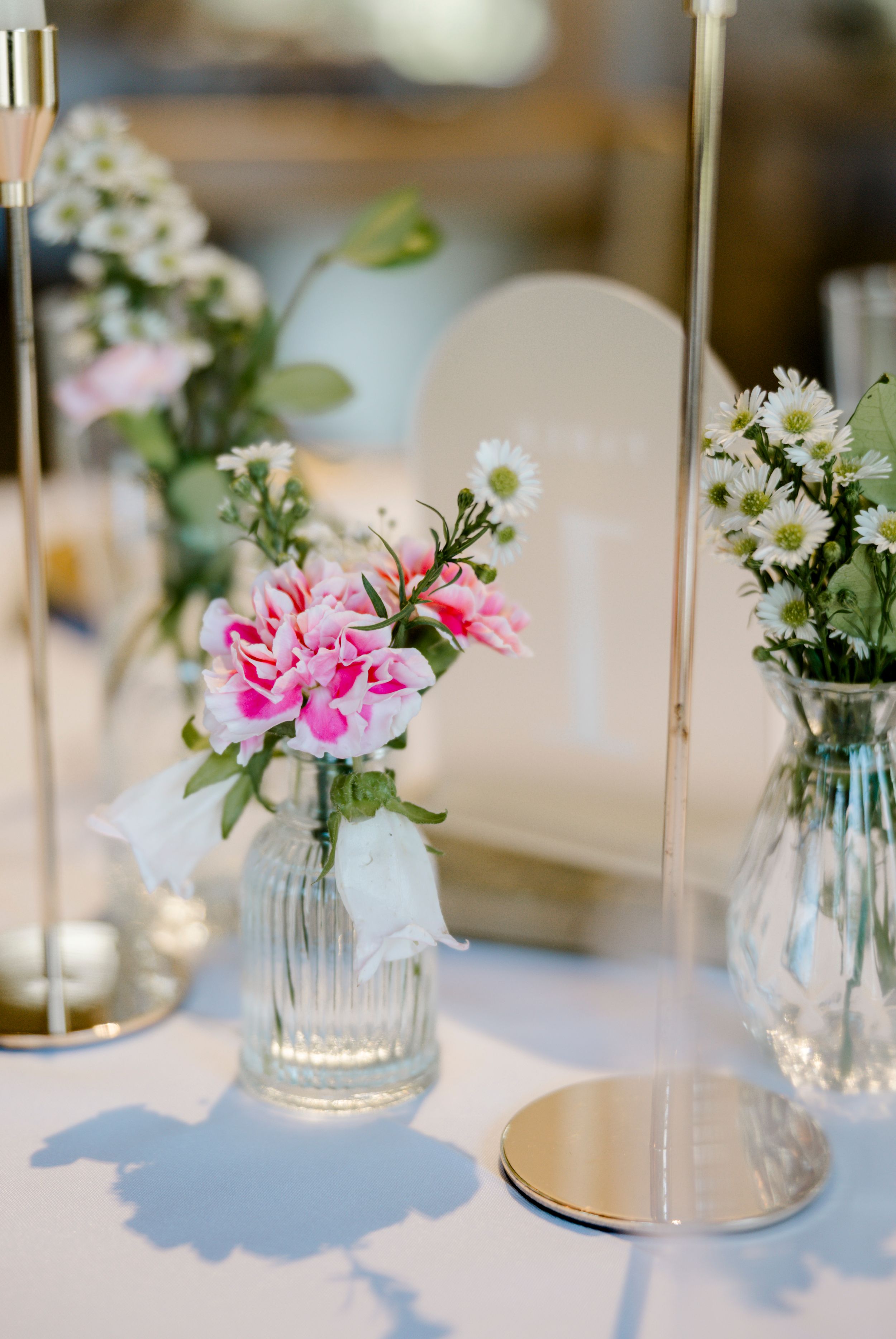 Small glass vases with pink carnations and white daisies create delicate centerpieces for an elegant table setting.