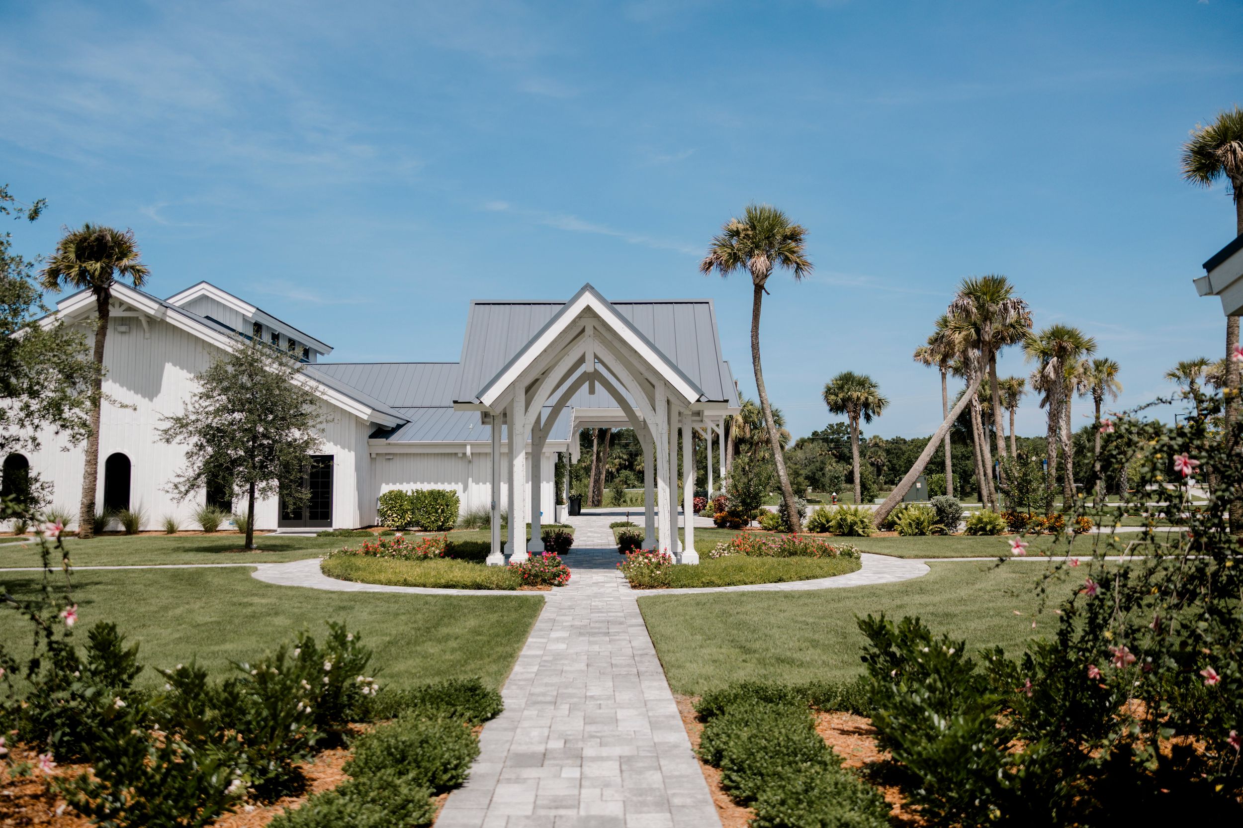 A scenic panoramic view of a white beachside chapel with palm trees and lush landscaping along a paved pathway.