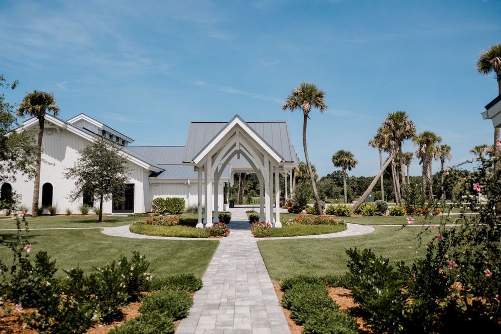 A scenic panoramic view of a white beachside chapel with palm trees and lush landscaping along a paved pathway.