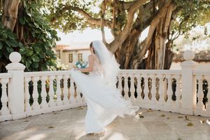 A couple in wedding attire shares a romantic moment on a terrace with ornate white stone balustrades and lush trees.