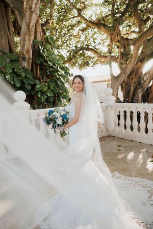Bride in flowing white wedding gown twirls gracefully under sunlit trees with veil floating in the breeze.