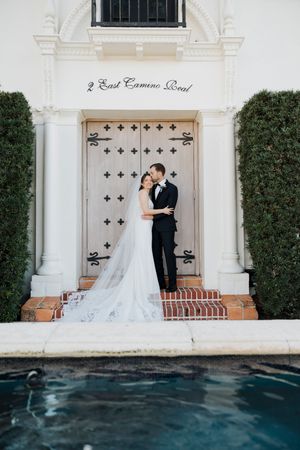 Elegant wedding couple poses by ornate white doors and columns near a swimming pool at dusk.