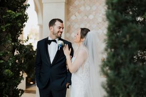 A joyful wedding couple shares an intimate moment outdoors with greenery and architecture in the background.