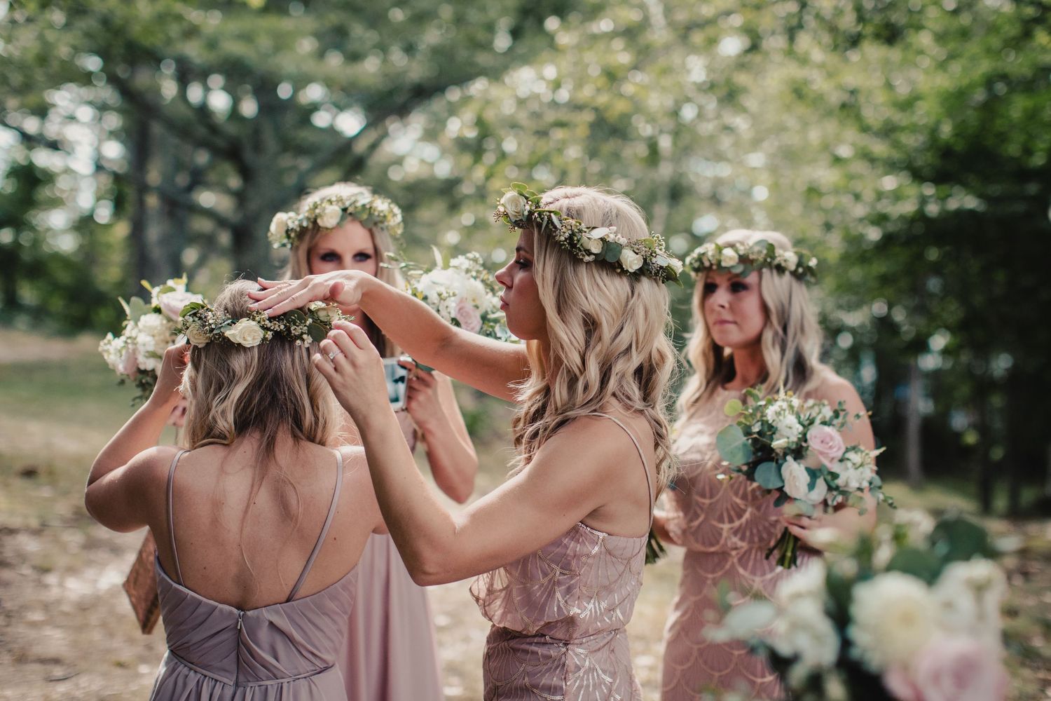 Bridesmaids in blush pink dresses adjusting floral crowns outdoors at a wedding ceremony.