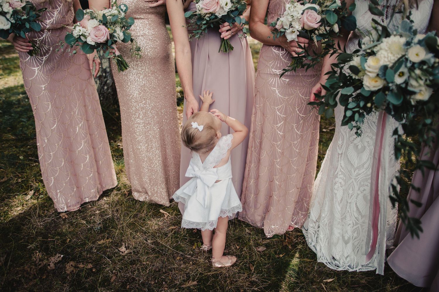 A toddler flower girl in white dress reaches up to bridesmaids in blush pink sequin gowns holding bouquets.