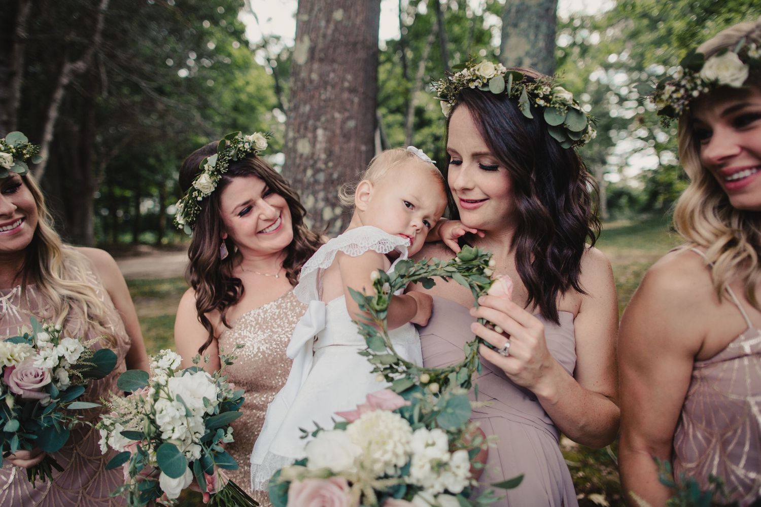 Bridesmaids in blush dresses with floral crowns hold bouquets while admiring a baby in a white outfit outdoors.