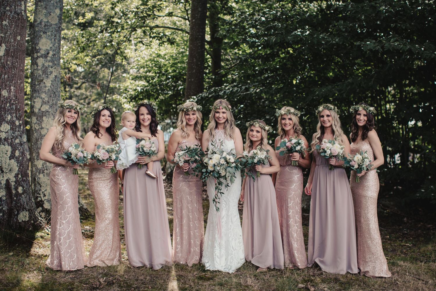 Bride and bridesmaids in blush gowns with floral crowns holding bouquets outdoors in a forest setting.