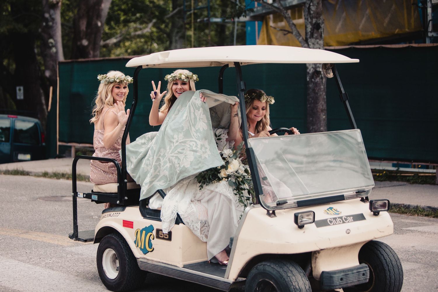 Bride and bridesmaids in floral crowns riding a golf cart outdoors, smiling and posing for wedding photos.