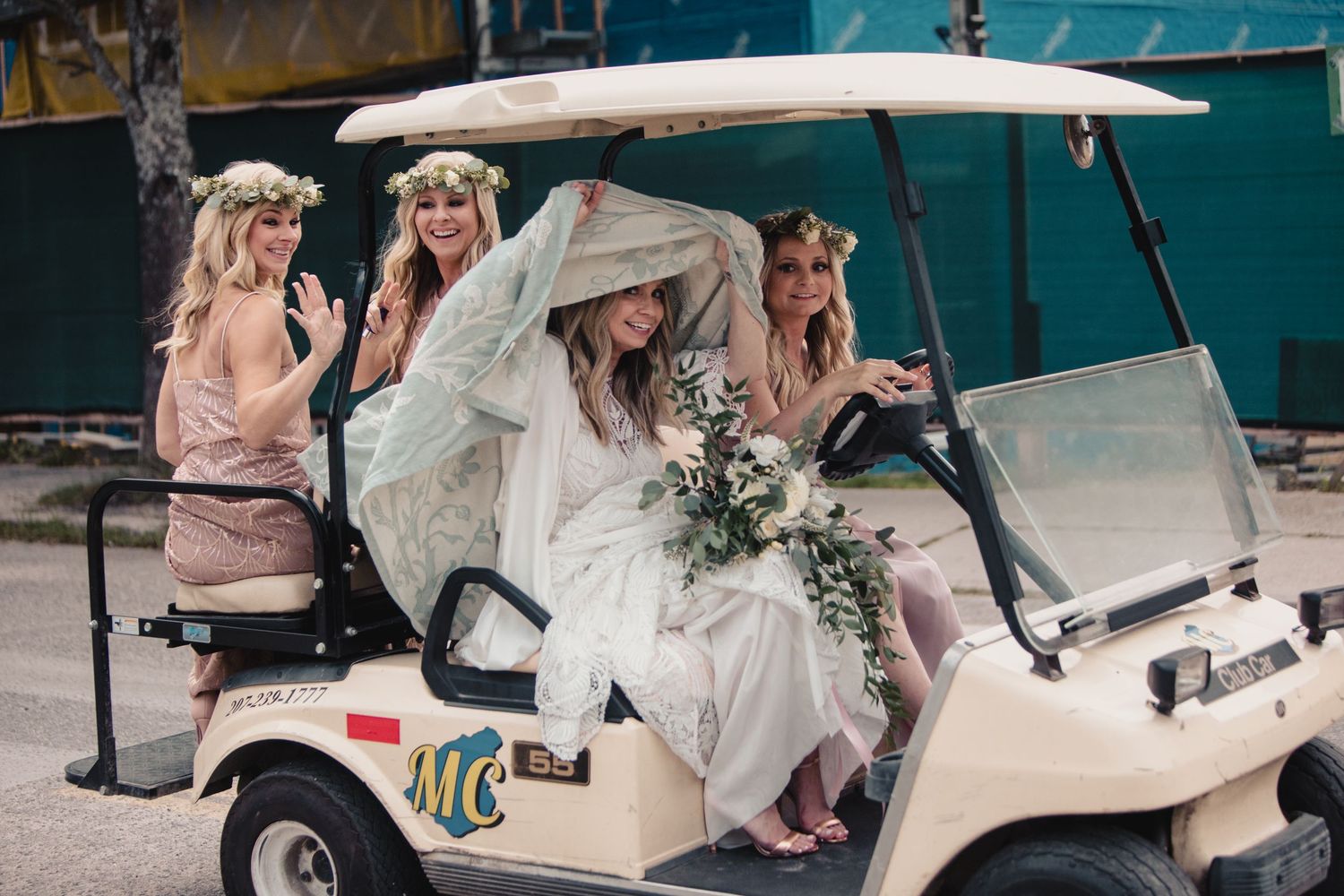 Bride and bridesmaids in floral crowns riding a golf cart decorated with greenery at an outdoor wedding.
