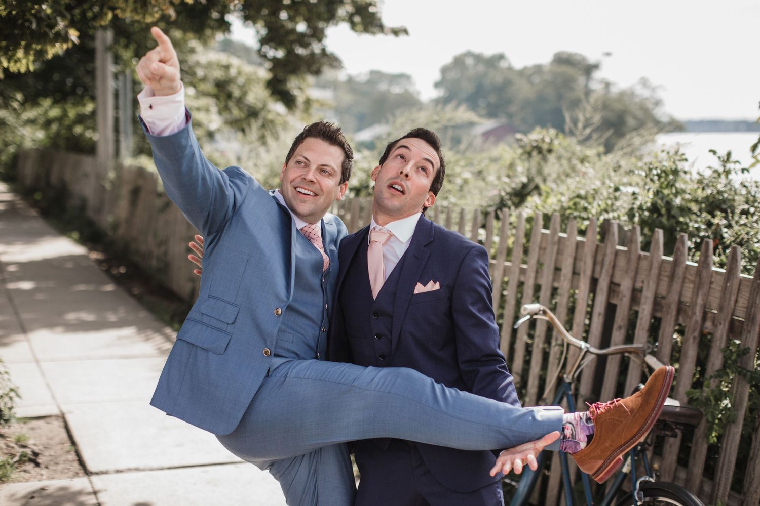 Two grooms in blue suits celebrate joyfully outdoors, one lifting the other as they pose near a fence on their wedding day.