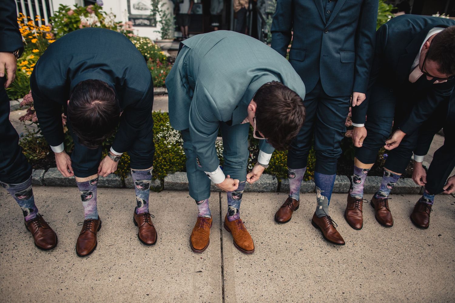 Groomsmen in teal suits showing off matching patterned socks and brown dress shoes on a sidewalk.