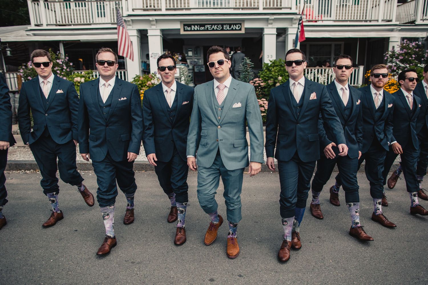 Groom in light gray suit leads groomsmen in navy suits and sunglasses walking outdoors at a wedding.