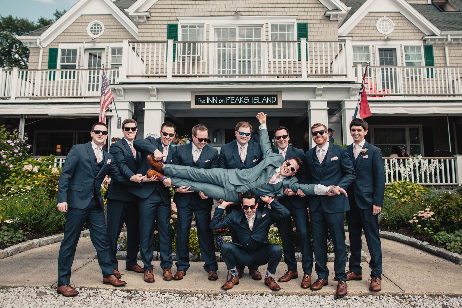 Groomsmen in navy suits lifting groom outside The Inn at Peaks Island during a fun wedding party photo.