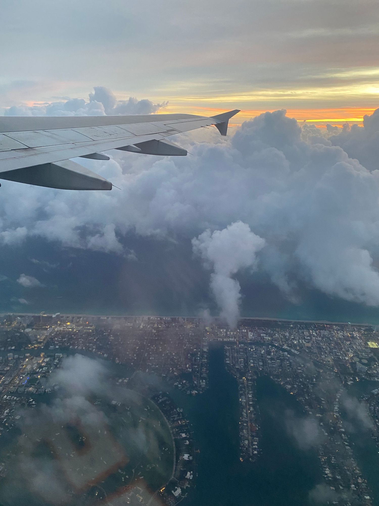 Aerial view of a city coastline at sunset with dramatic clouds from an airplane window.