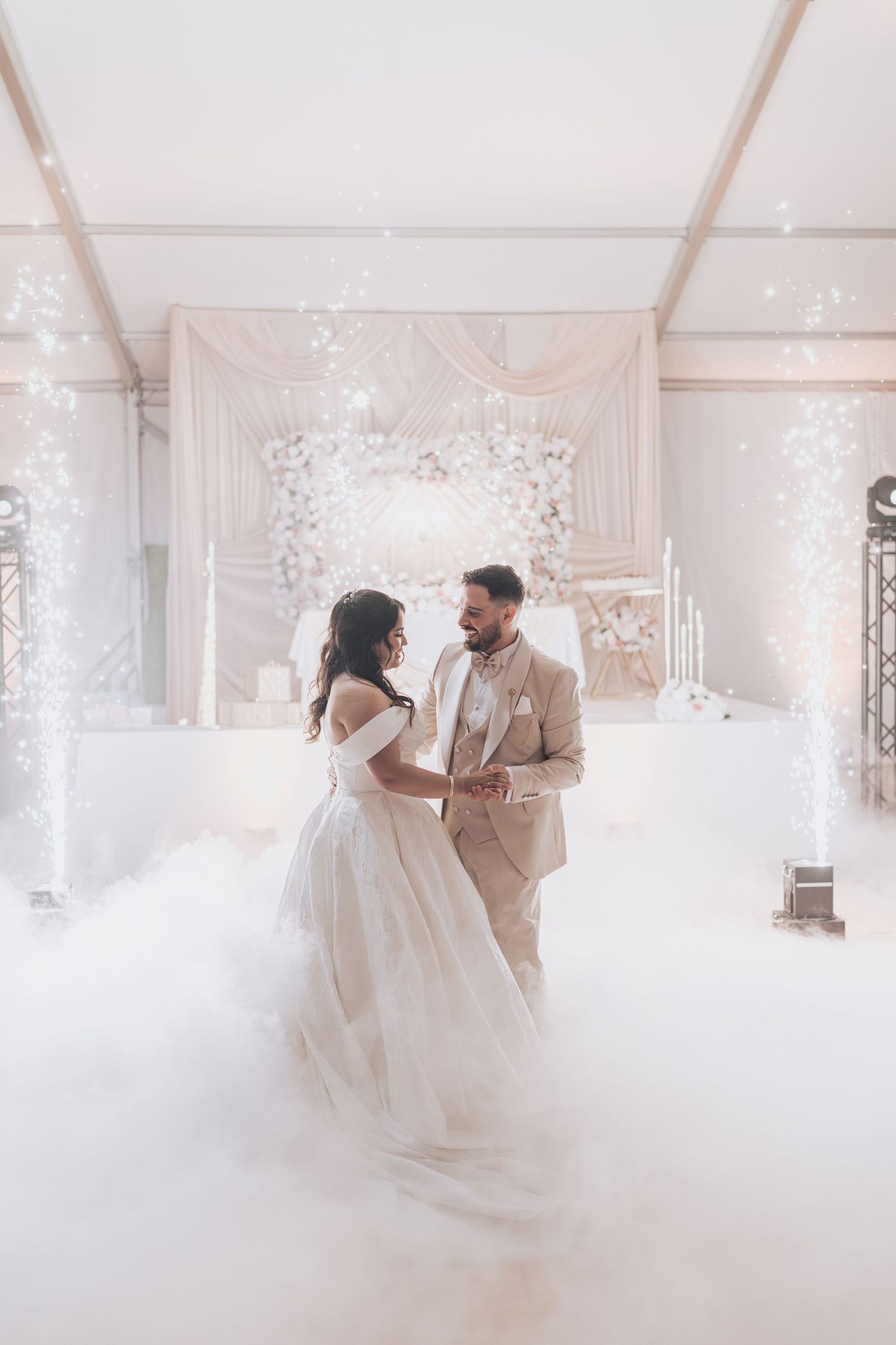 Bride and groom share first dance surrounded by dry ice fog and sparkling lights in elegant white wedding venue.