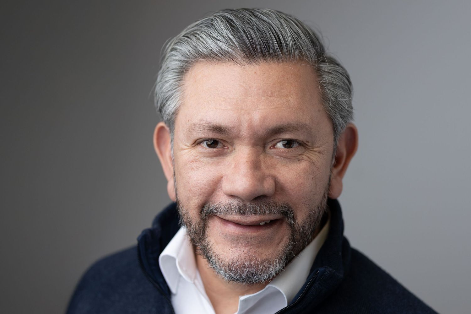 Smiling man with gray hair and beard wearing a navy sweater against a gray background.