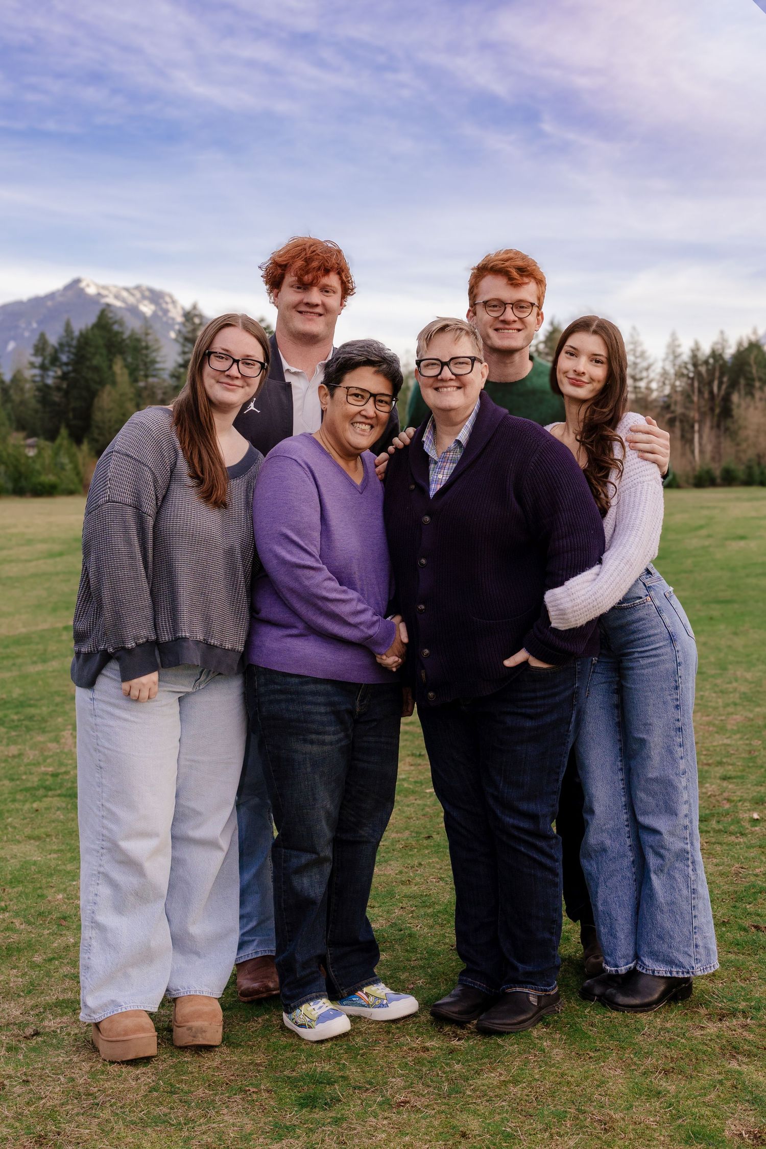 A group stands together smiling on a grassy field with snow-capped mountains and blue sky in the background.