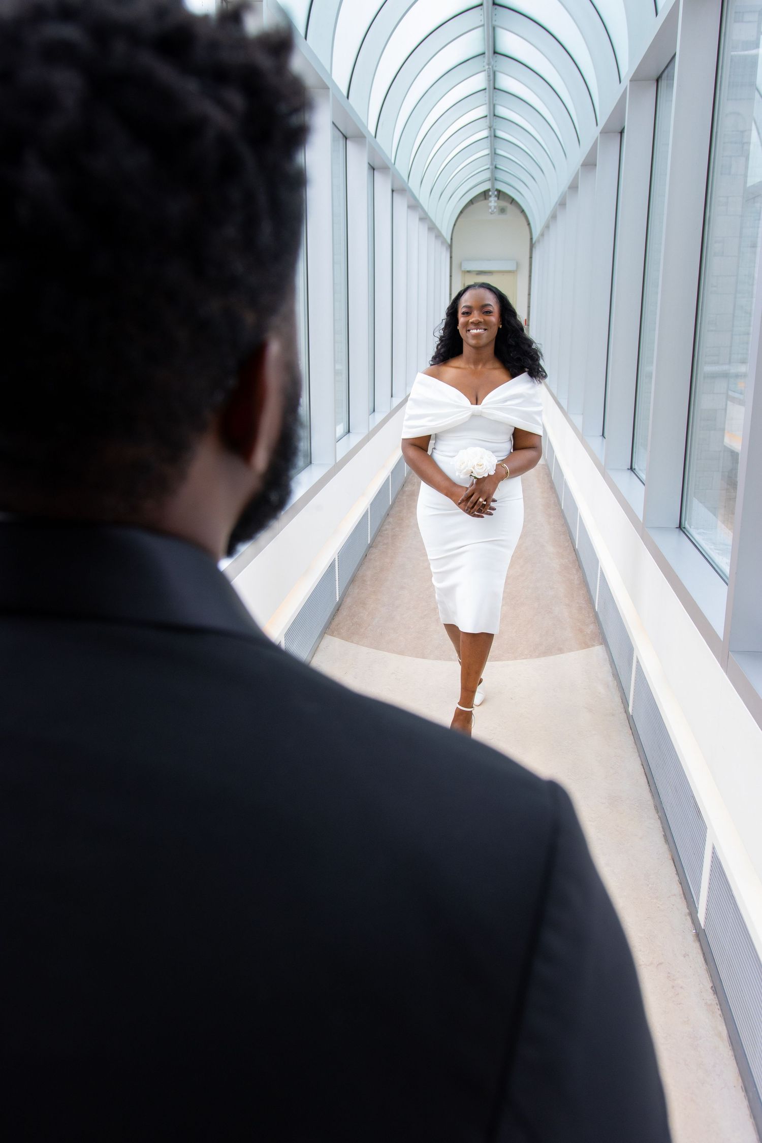 A bride in an elegant white off-shoulder dress walks down a bright, modern hallway with arched glass ceiling.