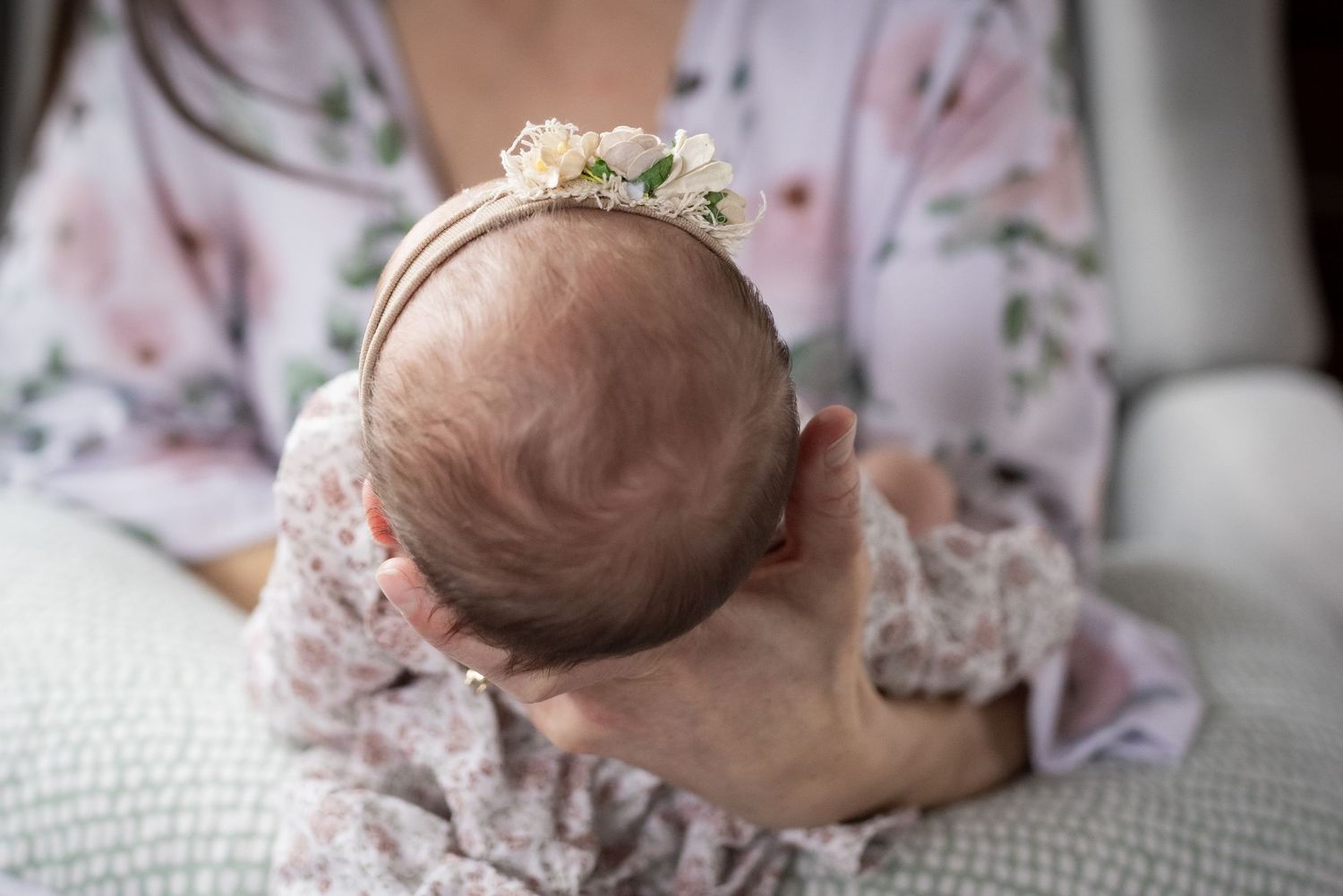 Mother cradles newborn baby head adorned with delicate floral headband from above.