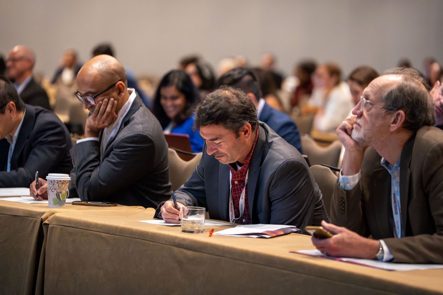 Business professionals seated at conference tables take notes and participate in a corporate meeting.