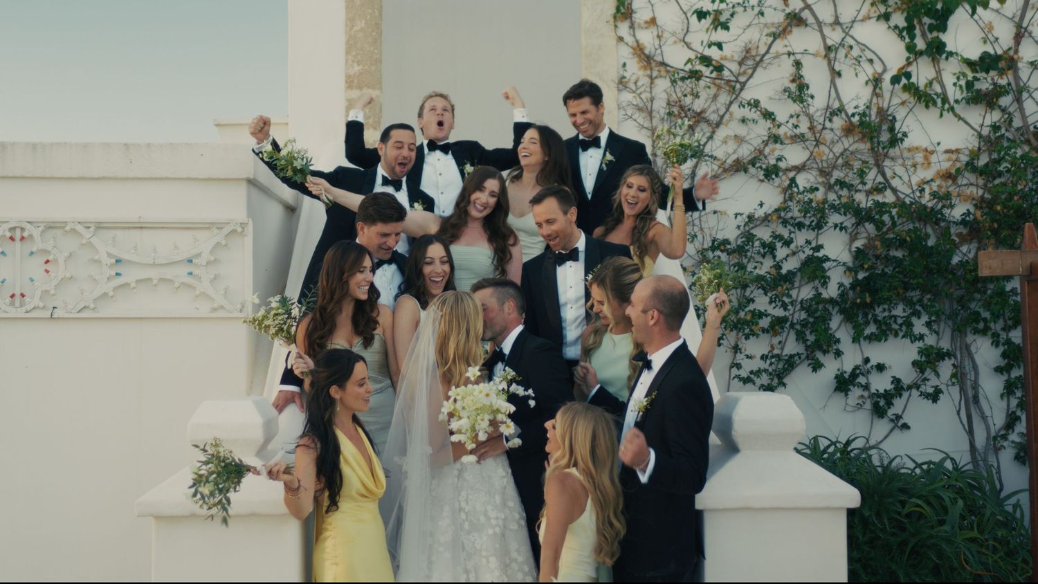 Wedding party poses on white marble stairs, bride holding bouquet surrounded by bridesmaids in yellow and green gowns and groomsmen.